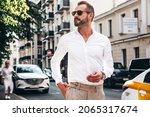 Portrait of handsome confident stylish hipster lambersexual model.Sexy modern man dressed in white shirt and trousers. Fashion male posing on street background in Europe city at sunset. In sunglasses