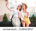 Two young beautiful smiling hipster female in trendy summer clothes.Sexy carefree women posing in the street. Positive pure models taking selfie. They drinking coffee or tea in plastic cup
