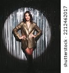 Small photo of Young beautiful smiling brunette female in trendy evening golden jacket. carefree woman posing near silver shiny tinsel wall in studio. Fashionable model with bright makeup looks at camera