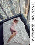 Small photo of woman in a nightgown lying on the bed window in a skyscraper near the bed