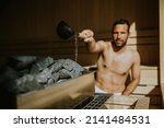 Handsome young man pouring water onto hot stone in the sauna