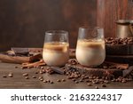 Irish cream and coffee cocktail in glasses with ice on an old wooden background. Coffee beans, cinnamon, anise, and pieces of chocolate are scattered on the table. 