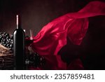 Small photo of Glass and bottle of red wine with grapes on a black reflective background. Red satin curtain flutters in the wind. Copy space.