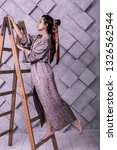 Small photo of Hairstyles look book. Prepossessing sophisticated with finely-cut face poser wearing elegant lilac maxi dress and beautified with scarf hairdo standing on ladder during studio shooting