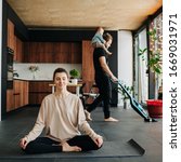 Small photo of Mother sits at easy simplified lotus pose. She's relaxed with her eyes closed, while father vacuum cleaning apartment floor with their infant baby riding on his neck