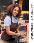 Small photo of Happy young mixed-race cashier with dark wavy hair looking at you while standing by workplace and pressing button of cashbox