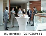 Large group of intercultural people in formalwear communicating at buffet while standing by tables served with snacks and champagne