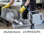 Small photo of Gloved hands of female cleaner throwing trash from garbage bin into plastic bucket on janitor trolley while working in modern office