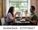 Small photo of Young woman and her smiling boyfriend with cups of coffee sitting by table in front of one another and chatting