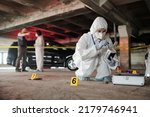 Small photo of Young criminological expert in coveralls inspecting empty bottle on crime scene while squatting in front of open briefcase with working supplies