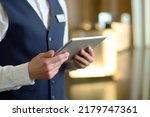 Small photo of Digital tablet in hands of young male receptionist of luxurious modern hotel standing in lounge by entrance and meeting new guests