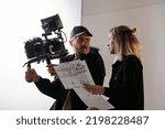Young bearded cameraman and his assistant with documents consulting about shooting while standing in front of camera in studio