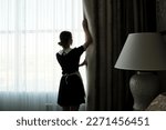 Back view of young chambermaid in uniform opening heavy night curtains in hotel room while standing in front of large window in the morning