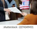 Close-up of young woman from electoral commission passing ballot paper to one of voters standing in queue in front of registration desk