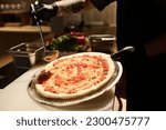 Chef with ladle spreading mixture of ingredients on surface of pizza flatbread while preparing it by workplace before baking it in oven