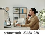 Side view of young confident chief executive officer or director of company talking to business partner on smartphone while sitting by workplace
