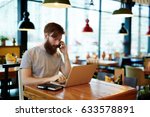 Busy bearded manager sitting in cyber cafe and talking to his business partner on mobile phone, modern laptop and notebook located on wooden table, waist-up portrait
