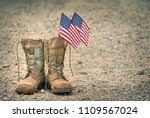 Small photo of Old military combat boots with dog tags and two small American flags. Rocky gravel background with copy space. Memorial Day or Veterans day concept.
