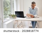 business man with blue shirt and black glasses is standing behind standing table and is working with his tablet and a black laptop
