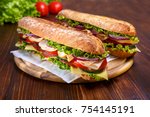 Two baguette sandwiches with salami, turkey breast, cheese, lettuce, tomatoes and onion on a cutting board. Long subway sandwiches on a dark background.