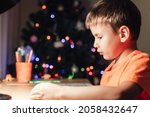 Small photo of 7 yeas old boy sitting at desk and reading book. Decorated Christmas tree on background