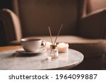 Liquid home fragrance with glass bottle and wooden sticks, cup of fresh tea staying on marble coffee table at home close up. Cozy atmosphere. 