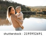 Smiling young woman holding playing with baby boy 1 year old wear knit clothes over nature background and lake with forest. Autumn season. Motherhood. 