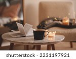 Cup of tea with paper open book and burning scented candles on marble table over cozy chair and glowing lights in bedroom closeup. Winter holiday season. 