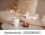 Coffee pot with cup of tea burning scented candle on marble table over Christmas lights in bedroom. Cozy home atmosphere. 