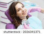 Small photo of Dentist choosing teeth enamel shade color for young smiling female patient before whitening procedure. Happy woman with beautiful white smile on stomatological appointment. Dentistry and stomatology.