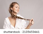 Small photo of Happy pretty young woman holding her long pigtail, pleased with healthy shining hairs posing on gray background. Beauty portrait of smiling girl with stylish braid. Haircare treatment products ad.