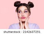 Small photo of Funny modern beautiful young girl with trendy lavender make up, pretty hairdo of two hair buns looking aside, touching cheeks, pouted lips. Stylish pensive teen with lilac eyeshadow feel puzzlement.