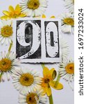 Small photo of The number ninety stamped on paper and surrounded by pressed daisies and winter aconite flowers . A yellow, white , green and black floral ninetieth birthday card image