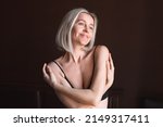 Small photo of Attractive middle-aged woman enjoys herself and her body, loves herself. 40-year-old female hugs herself in the bedroom at home in the morning