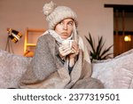 Small photo of Frozen. Sad woman sit on couch at freezing cooled house in warm cap and blanket shiver tremble with cold. Unhappy middle aged lady spend time at home feel bad suffer of heating system problems