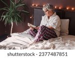 Refreshed elderly 60s female wakes up in morning, reading book, seated in bed in bedroom at home, middle-aged woman feels happy and peppy after enough sleeping, greeting new day, relaxing concept
