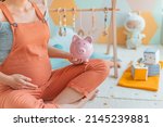Pregnancy family planning budget. Cost of having a child. Pregnant woman holding piggy bank shopping newborn toys and nursery decor with savings. Maternity leave benefits