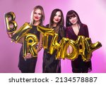 Three women holding Golden HAPPY BIRTHDAY words made of inflatable balloons in hands on pink background