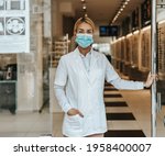 Young and attractive female optometrist with face protective mask standing at open optical store doors and looking outside. She is confident and serious. Covid-19 open for business concept.