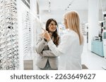Beautiful and fashionable woman choosing eyeglasses frame in modern optical store. Female seller specialist helps her to make right decision.