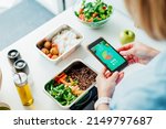 Healthy diet plan for weight loss, daily ready meal menu. Woman using meal tracker app on phone while weighing lunch box cooked in advance on kitchen scale. Balanced portion with dish. Pre-cooking
