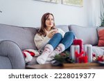 Positive woman in plaid with cup of tea watching movie, TV on sofa at home with christmas decoration atmosphere. Lady wear jumper and warm socks. Cozy and comfortable winter concept. Selective focus.