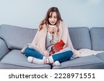 Woman freezes in wintertime. Young girl wearing warm woolen socks and wrapped into two blankets, holding a cup of hot drink and heating pad while sitting on sofa at home. Keep warm. Selective focus.