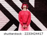 Hipster fashion young woman in trendy magenta color sweatshirt and sunglasses and bucket hat posing on the painted brick wall background. Color of the 2023 year. Urban city street fashion.