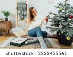Young woman in cozy sweater decorating potted Christmas tree with small glass baubles in light modern Scandinavian interior. Eco-friendly winter holidays. Christmas tree in a pot. Selective focus