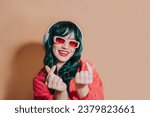 Small photo of Cute kawaii girl with green hair and headphones, red lips, sunglasses showing korean hearts with fingers and smiling, laughing silly, posing on beige background happy and joyful, K pop culture fans.