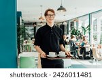 Small photo of Portrait of young smiling affable waiter carrying on tray with coffee cups to client table in cafe. Hospitality service job. Occupation and work concept. Part-time work for students. Selective focus