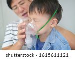 Asian father helping his toddler son with inhalation therapy by the mask of inhaler. Sick little kid with respiratory problem with oxygen mask breathes through nebulizer at doctor clinic