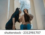 Small photo of Low angle view shot of two lovely girls listening to same song on different headphones. Brunette girl in black overcoat singing to her friend